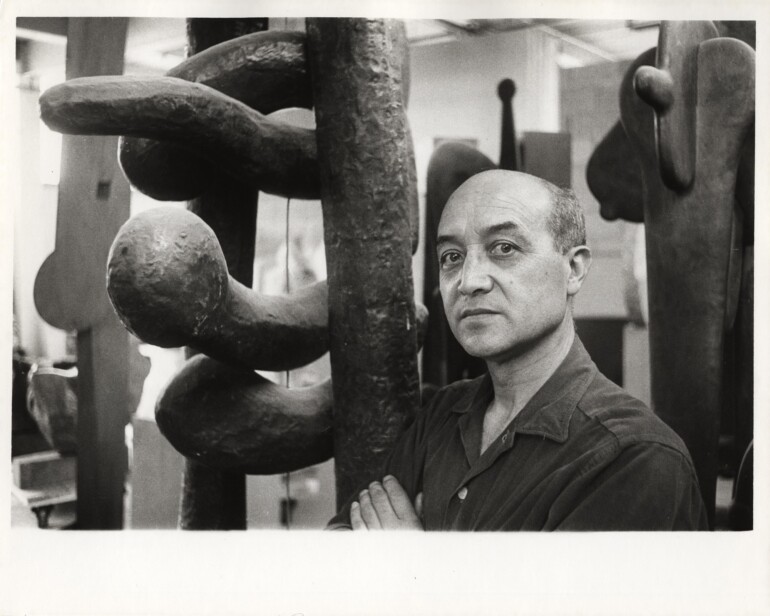 Portrait of artist and designer Isamu Noguchi standing next to one of his sculptures